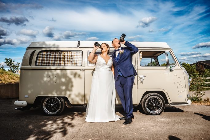 Braut und Bräutigam lehnen an einem Auto, trinken gemeinsam Champagner