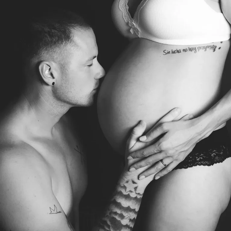 Mann mit freiem Oberkörper küsst zärtlich den Babybauch seiner Frau.